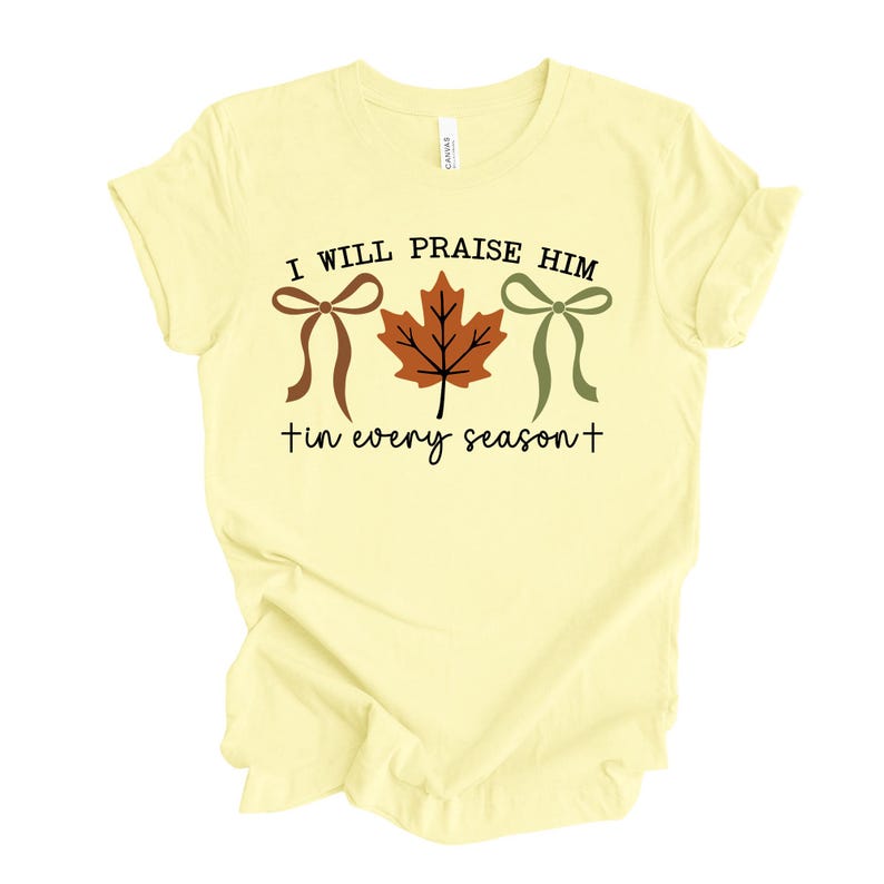 May include: A light yellow t-shirt with the text "I WILL PRAISE HIM in every season" in black. The design includes a brown bow, a brown and orange leaf, and a green bow.