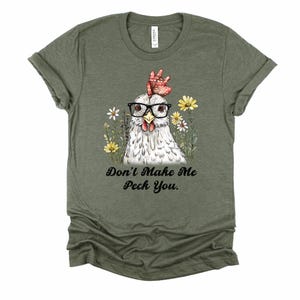 May include: Olive green t-shirt featuring a whimsical illustration of a chicken wearing glasses and surrounded by flowers. The text "Don't Make Me Peck You" is printed below the chicken. The shirt is made of a soft material.