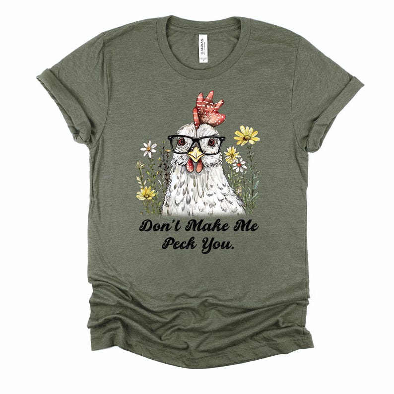 May include: Olive green t-shirt featuring a whimsical illustration of a chicken wearing glasses and surrounded by flowers. The text "Don't Make Me Peck You" is printed below the chicken. The shirt is made of a soft material.
