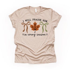 May include: Beige t-shirt with the text "I WILL PRAISE HIM + in every season +." The design includes a brown bow, an orange leaf, and a green bow. The shirt is a short-sleeved crew neck.
