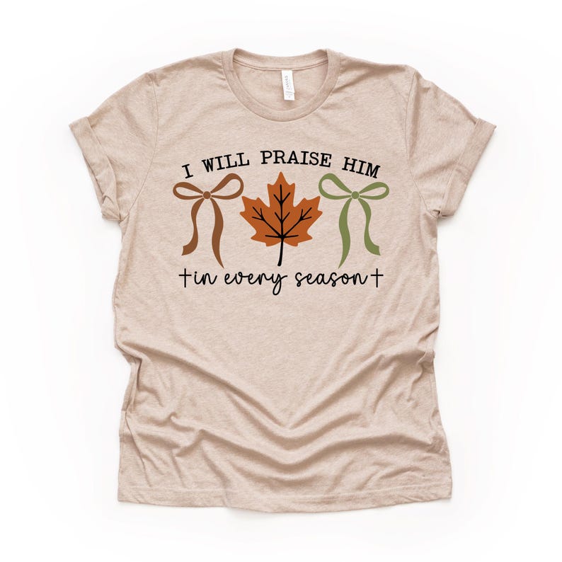 May include: Beige t-shirt with the text "I WILL PRAISE HIM + in every season +." The design includes a brown bow, an orange leaf, and a green bow. The shirt is a short-sleeved crew neck.