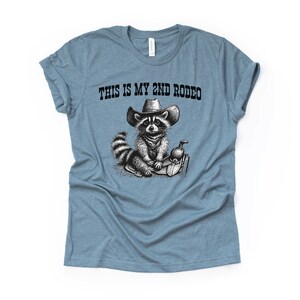 Pode incluir: Uma camiseta azul claro com uma ilustra&ccedil;&atilde;o em preto e branco de um guaxinim usando um chap&eacute;u de cowboy e montando uma sela. O texto "This is my 2nd Rodeo" est&aacute; impresso na camiseta.