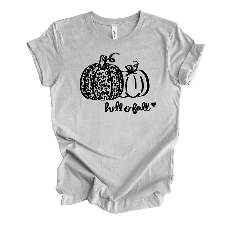 May include: A light gray t-shirt featuring a black graphic of two pumpkins. One pumpkin has a leopard print design, and the other is solid. The words "hello fall" are written below the pumpkins, with a heart symbol.