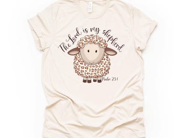 Könnte beinhalten: Ein beiges T-Shirt mit einem braunen und weißen Leopardenmuster-Cartoon-Schaf. Der Text "The Lord is my shepherd" ist in einer kursivschriftlichen Schriftart über dem Schaf geschrieben. Der Text "Psalm 23:1" ist unter dem Schaf geschrieben.