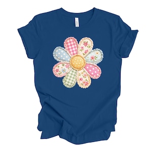 May include: A blue t-shirt featuring a large, appliqued flower design. The flower has a yellow center and petals in various patterns, including pink polka dots, floral prints, and gingham. The shirt is made of a soft material.