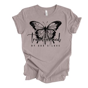 Peut inclure: Un t-shirt gris chin&eacute; avec un motif de papillon noir et le texte "transform&eacute; par l'amour de Dieu".