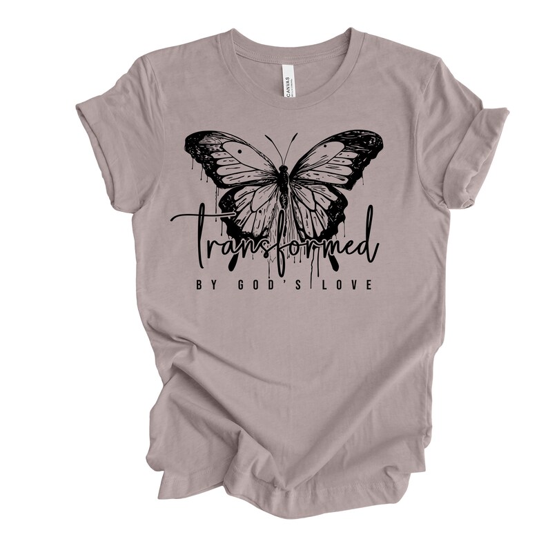 Peut inclure: Un t-shirt gris chin&eacute; avec un motif de papillon noir et le texte "transform&eacute; par l'amour de Dieu".