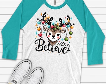 May include: A white and turquoise raglan t-shirt with a cartoon reindeer wearing Christmas lights and the word "Believe" printed on the front. The shirt is paired with white canvas sneakers.