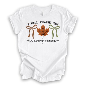 May include: White t-shirt with the text "I WILL PRAISE HIM" above a brown bow, a brown maple leaf, and a green bow. Below the graphic is the text "tin every season+". The shirt is made of a soft material.