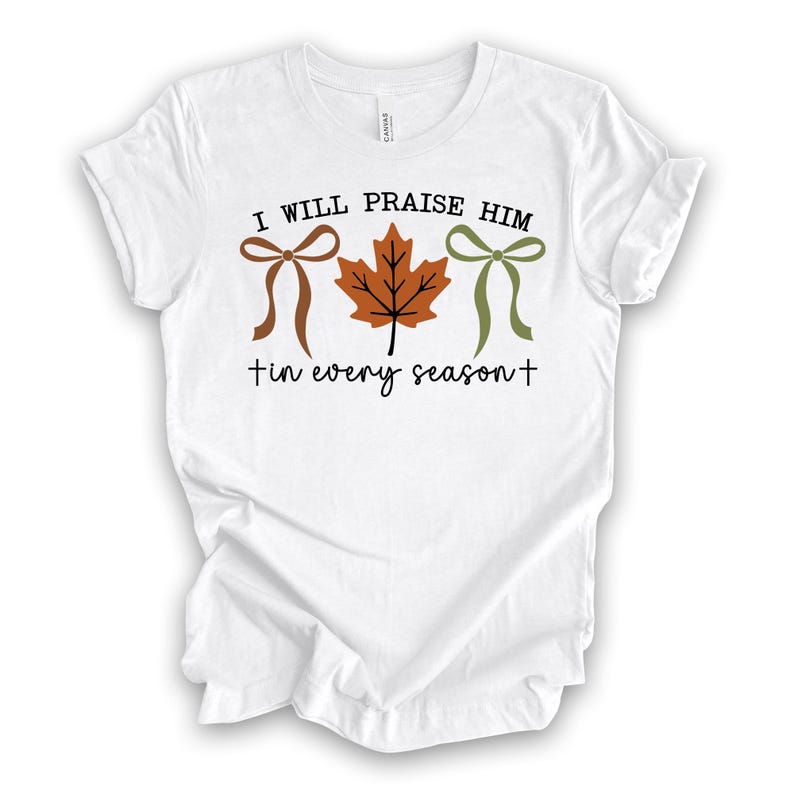 May include: White t-shirt with the text "I WILL PRAISE HIM" above a brown bow, a brown maple leaf, and a green bow. Below the graphic is the text "tin every season+". The shirt is made of a soft material.