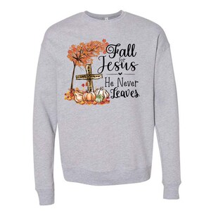 Puede incluir: Una sudadera gris con un dise&ntilde;o de oto&ntilde;o que presenta una cruz, calabazas y hojas. El texto dice "Fall for Jesus He Never Leaves."