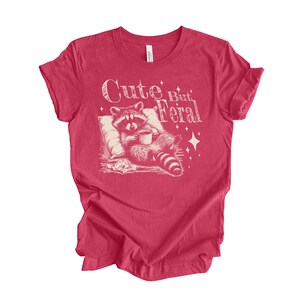 May include: Red t-shirt with a white graphic of a raccoon holding a cup of coffee and the text "Cute But Feral".