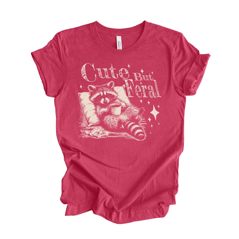 May include: Red t-shirt with a white graphic of a raccoon holding a cup of coffee and the text "Cute But Feral".