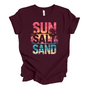 Op de afbeelding: Een bordeauxrood t-shirt met de woorden "SUN SALT SAND" in een kleurrijk, gelaagd ontwerp. Het ontwerp bevat een zonsondergang, palmbomen en oceaangolven. Het shirt is gemaakt van een zacht materiaal.