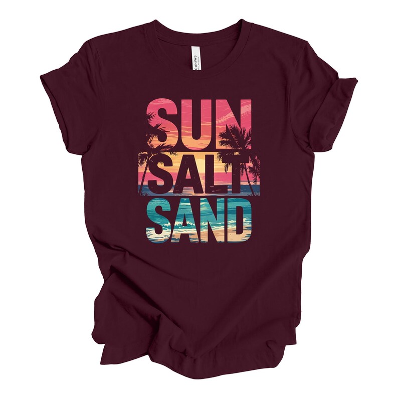 Op de afbeelding: Een bordeauxrood t-shirt met de woorden "SUN SALT SAND" in een kleurrijk, gelaagd ontwerp. Het ontwerp bevat een zonsondergang, palmbomen en oceaangolven. Het shirt is gemaakt van een zacht materiaal.