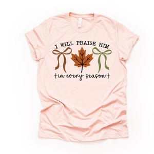 May include: A light pink t-shirt with the text "I WILL PRAISE HIM" above a brown bow, a brown maple leaf, and a green bow. Below the graphic is the text "tin every season+".