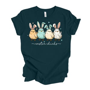 Könnte beinhalten: Dunkelblaues T-Shirt mit einer Grafik von vier Cartoon-Küken mit Hasenohren. Die Küken sind in Gelb-, Blau- und Weißtönen gehalten. Unter den Küken steht in Schreibschrift "easter chicks".