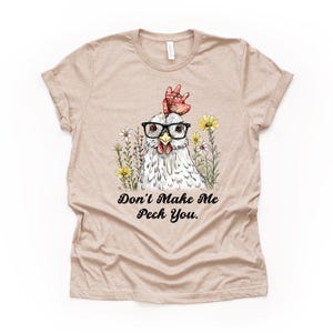 May include: A tan t-shirt featuring a whimsical illustration of a chicken wearing glasses and surrounded by flowers. The text "Don't Make Me Peck You" is printed below the chicken. The shirt has short sleeves and a crew neck.