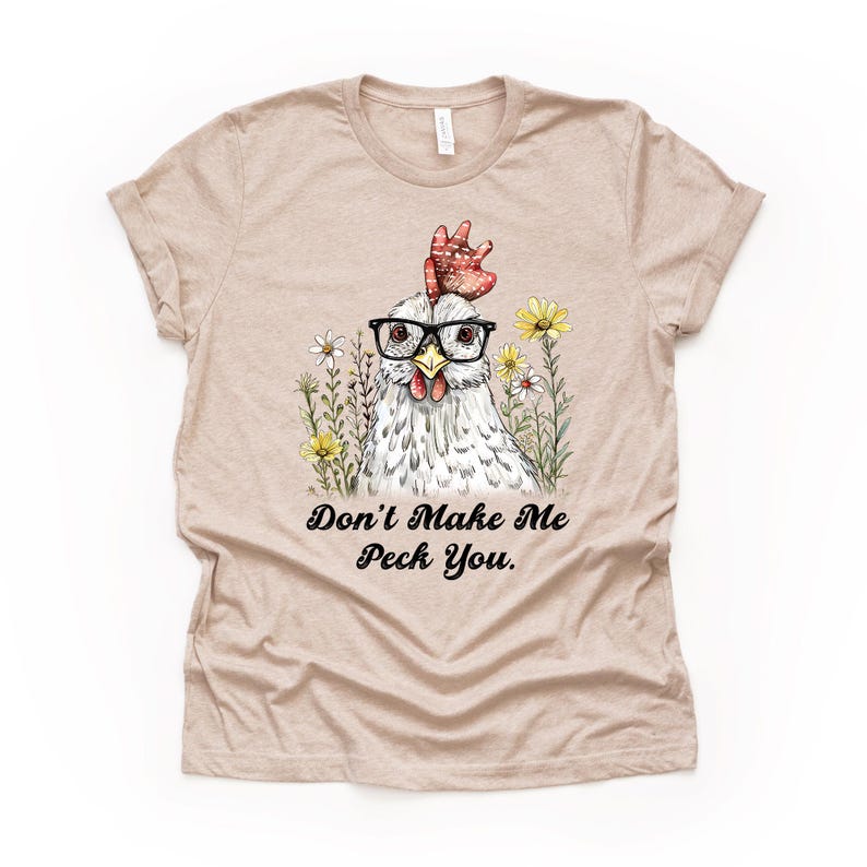 May include: A tan t-shirt featuring a whimsical illustration of a chicken wearing glasses and surrounded by flowers. The text "Don't Make Me Peck You" is printed below the chicken. The shirt has short sleeves and a crew neck.