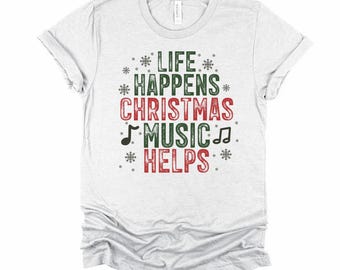 May include: A white t-shirt with the text "LIFE HAPPENS CHRISTMAS MUSIC HELPS" in red and green, with musical notes and snowflake accents. The shirt is made of a soft material and has a classic crew neck.
