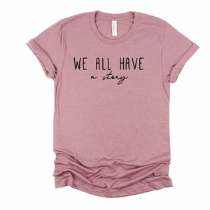 May include: A mauve coloured t-shirt with the text "WE ALL HAVE a story" printed in black.