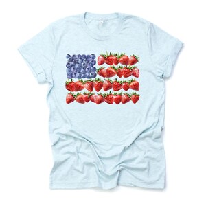 May include: Light blue t-shirt featuring a unique design of the American flag made from blueberries and strawberries. The flag design is centered on the front of the shirt. The shirt is made of a soft, lightweight material.