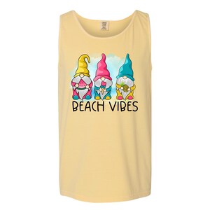 Pu&ograve; includere: Una canotta gialla con una grafica di tre gnomi che tengono dolcetti estivi. Gli gnomi indossano cappelli colorati e sono in piedi davanti a un cielo blu e bianco. Il testo "Beach Vibes" &egrave; stampato sotto gli gnomi.