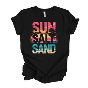 Op de afbeelding: Zwart t-shirt met de tekst "SUN SALT SAND" in een kleurrijk strandontwerp. De tekst is gelaagd met een zonsondergang en palmbomen, wat een tropische vakantiesfeer oproept.