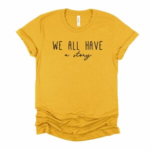 May include: A mustard yellow t-shirt with the text "WE ALL HAVE a story" printed in black.