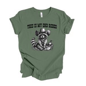 Pode incluir: Camiseta verde com uma ilustra&ccedil;&atilde;o em preto e branco de um guaxinim usando um chap&eacute;u de cowboy e sentado em uma sela. O texto "This is my 2nd Rodeo" est&aacute; impresso na camiseta.