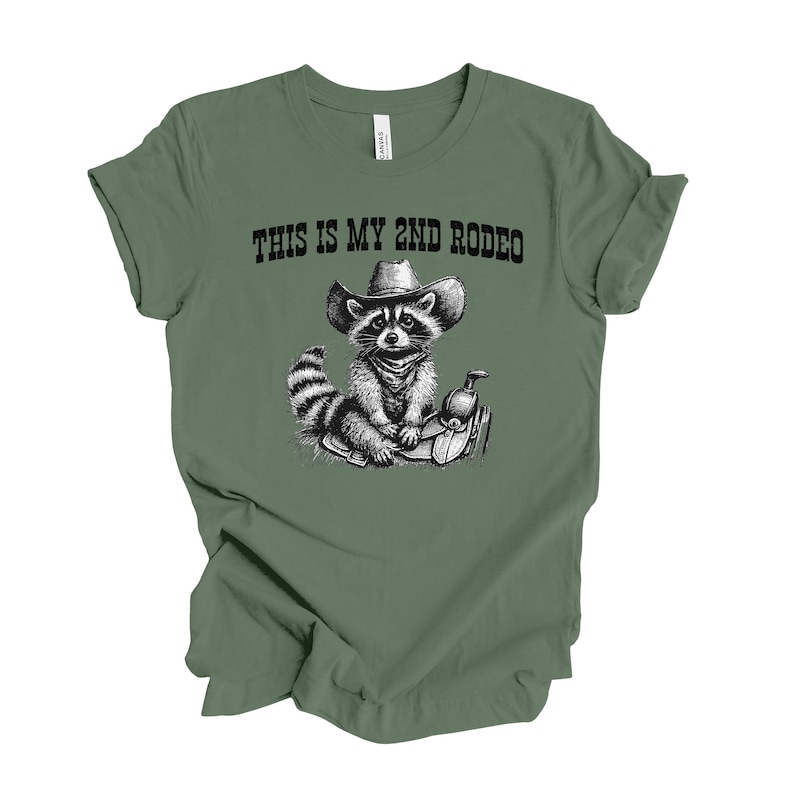 Pode incluir: Camiseta verde com uma ilustra&ccedil;&atilde;o em preto e branco de um guaxinim usando um chap&eacute;u de cowboy e sentado em uma sela. O texto "This is my 2nd Rodeo" est&aacute; impresso na camiseta.