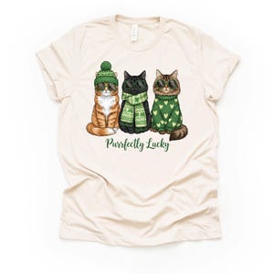 May include: Cream-colored t-shirt featuring three cartoon cats. The cats are dressed in St. Patrick's Day attire, including a green hat, sunglasses, and sweaters. The text "Purrfectly Lucky" is printed below the cats.