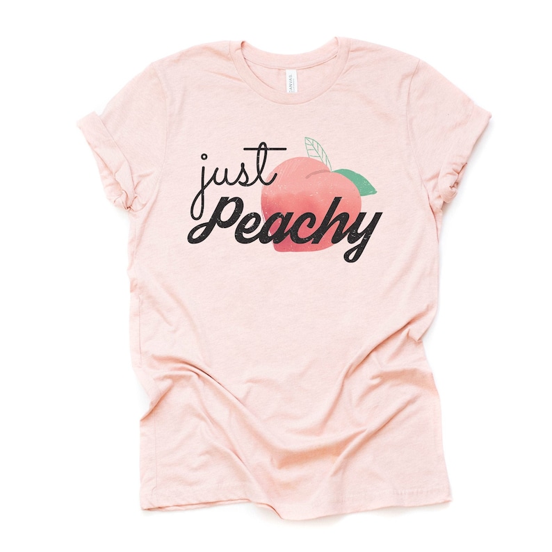 Może przedstawiać: R&oacute;żowy T-shirt z napisem "just peachy" w czarnym, ozdobnym piśmie. Obok napisu znajduje się brzoskwinia z zielonym liściem.