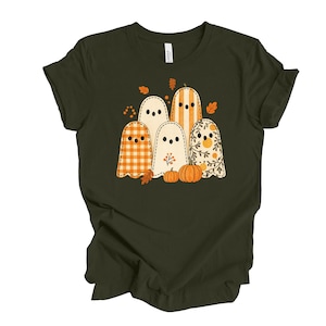 May include: Dark green t-shirt featuring a graphic of five cartoon ghosts with various patterns, including stripes and plaid, and two small pumpkins. The ghosts are surrounded by autumn leaves, creating a fall or Halloween theme.