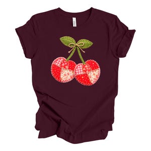 May include: Burgundy t-shirt featuring a whimsical cherry design. The cherries are crafted from patchwork fabric in red, pink, and white patterns, with green leaves and a brown stem. The design is centered on the front of the shirt.