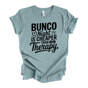 K&ouml;nnte beinhalten: Hellblaues T-Shirt mit dem schwarzen Text "Bunco Night is Cheaper Than Therapy". Der Text enth&auml;lt ein Herz, W&uuml;rfel und Ausrufezeichen. Das T-Shirt ist aus weichem Stoff.