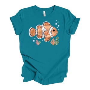 May include: A teal short-sleeve t-shirt featuring a central graphic of a clownfish with an orange and white gingham pattern, outlined in dark stitching. The design includes small blue bubbles and colorful coral sprigs below the fish.