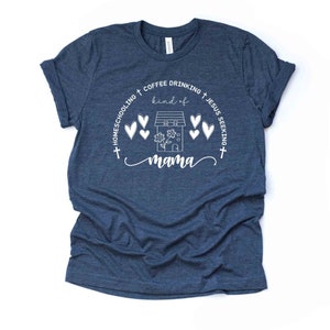 Puede incluir: Una camiseta azul marino con un dise&ntilde;o gr&aacute;fico blanco que dice "Homeschooling + Coffee Drinking + Jesus Seeking + kind of mama" con una casa y corazones alrededor.