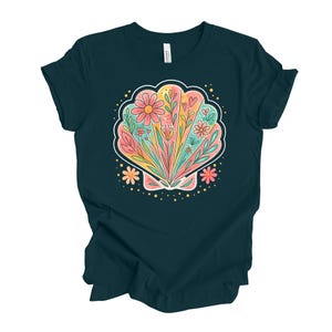 May include: A dark teal t-shirt featuring a colorful seashell design. The seashell is filled with flowers, leaves, and hearts in shades of pink, yellow, and orange. The design is outlined in white and dotted with small gold circles.