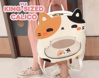 Puede incluir: Mochila con forma de gato calicó blanco, negro y naranja con el texto "V1.5 KING-SIZED CALICO". La mochila tiene una ventana transparente y correas ajustables. La bolsa tiene un asa superior y herrajes dorados.