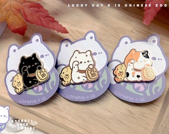 Könnte beinhalten: Drei Emaille-Anstecknadeln mit Glückskatzen-Designs, jeweils auf einer lila Rückkarte. Die Katzen sind schwarz, weiß und Schildpattfarben und halten Goldmünzen. Der Text "LUCKY CAT X 12 CHINESE ZODIAC" steht oben.