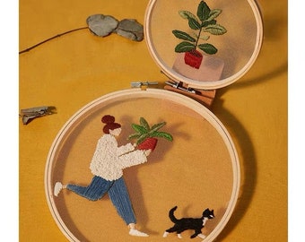 May include: Two embroidery hoops with botanical themes. One hoop features a plant in a red pot, while the other depicts a person carrying a plant, followed by a black and white cat. The hoops are set against a yellow background.