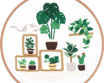 Può includere: Ricamo su telaio raffigurante varie piante in vaso e un gatto. Le piante includono una monstera, un cactus con fiori rossi e altro verde. Il ricamo è in un telaio di legno.