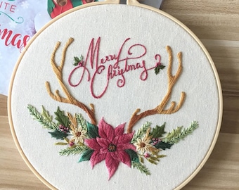 May include: Embroidered hoop art with the words "Merry Christmas" in red script. The design includes brown antlers, a pink poinsettia, and green foliage. The embroidery is set in a wooden hoop.