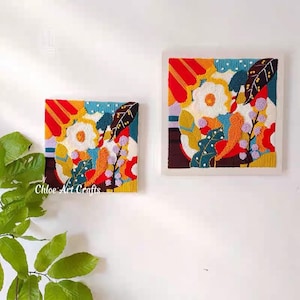 May include: Two framed needlepoint embroidery art pieces with a colorful abstract floral design. The larger piece is on the top and the smaller piece is on the bottom. Both pieces have a white background and a white frame.