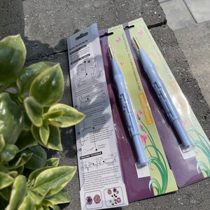 May include: Two punch needle tools in packaging. The tools are light blue with gold tips. The packaging has instructions and floral designs. The text on the packaging reads "Needle Punching is easy to start and impossible to stop!" and "Quality Product".