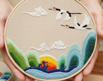 Può includere: Opera d'arte ricamata in un telaio di legno raffigurante un paesaggio con montagne, un sole al tramonto e gru in volo. L'opera utilizza sfumature di verde, blu, arancione e bianco su uno sfondo beige. Il telaio è tenuto da mani.