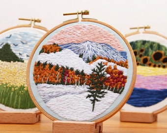Può includere: Arte paesaggistica ricamata in cerchi di legno. Il cerchio centrale presenta una scena di montagna innevata con alberi arancioni e rossi, un pino verde e un cielo rosa e blu. Altri cerchi raffigurano un campo di fiori gialli e girasoli.