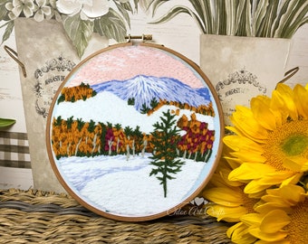 Può includere: Ricamo fatto a mano in un telaio di legno. L'opera raffigura un paesaggio montano invernale con terreno innevato, alberi colorati e una catena montuosa blu sotto un cielo rosa e blu. Il nome dell'artista, "Chloe Art Crafts", è visibile.