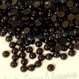 May include: A large quantity of black round plastic cabochons. The cabochons are smooth and shiny. They are scattered on a white surface.